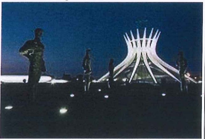 Fotografia noturna da Catedral Metropolitana de Brasília, iluminada, com estátuas em silhueta em primeiro plano.