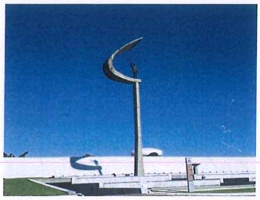 Fotografia do Memorial JK, com o pedestal alto encimado pela estátua de Juscelino sob uma estrutura em forma de arco, e o edifício branco ao fundo.