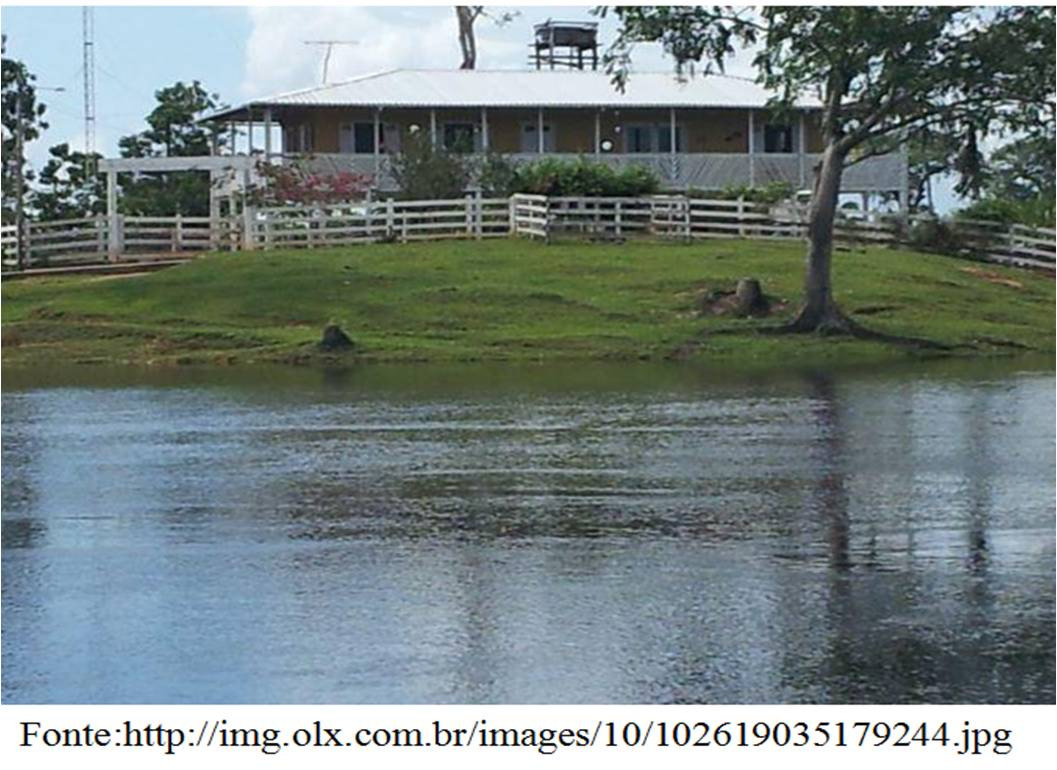Fotografia de uma casa de sítio com varanda, cercada por cerca de madeira branca, sobre um gramado à beira de um rio ou lago; há árvores ao redor e uma caixa-d'água no telhado.