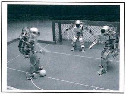Fotografia de três robôs humanoides jogando futebol em uma quadra, um deles conduzindo a bola diante de um gol.