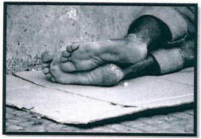 Fotografia em preto e branco dos pés descalços e sujos de uma pessoa deitada sobre um papelão na calçada.