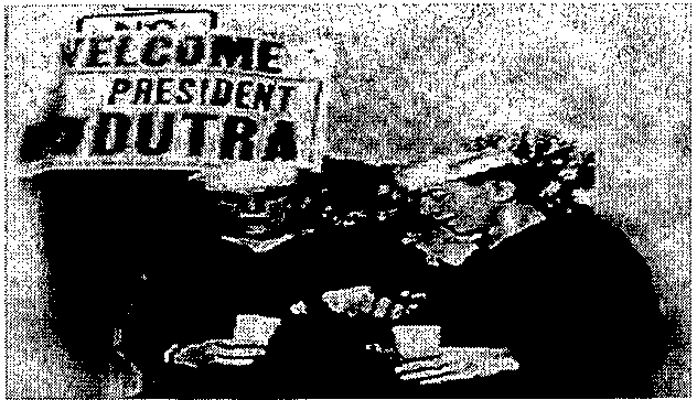 Fotografia: faixa com os dizeres “WELCOME PRESIDENT DUTRA” acima de pessoas reunidas.