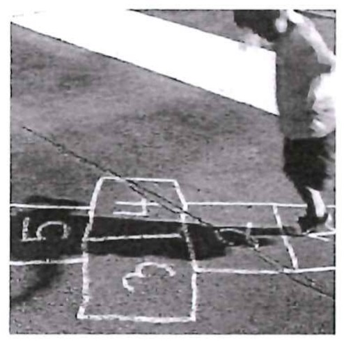 Fotografia em preto e branco de uma criança pulando amarelinha desenhada a giz no chão, com casas numeradas.