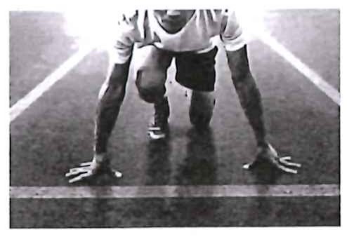 Fotografia em preto e branco de um atleta agachado na posição de largada, com as mãos apoiadas na pista.