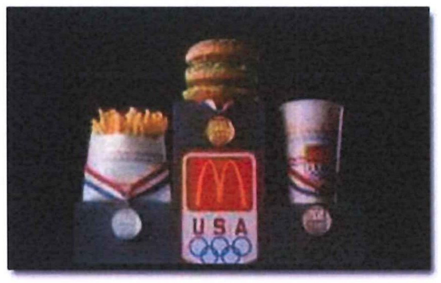 Fotografia de embalagens promocionais do McDonald’s sobre fundo escuro: batata-frita, sanduíche Big Mac e copo de refrigerante, com medalhas penduradas e um painel com o logotipo da rede, a inscrição “USA” e os anéis olímpicos.