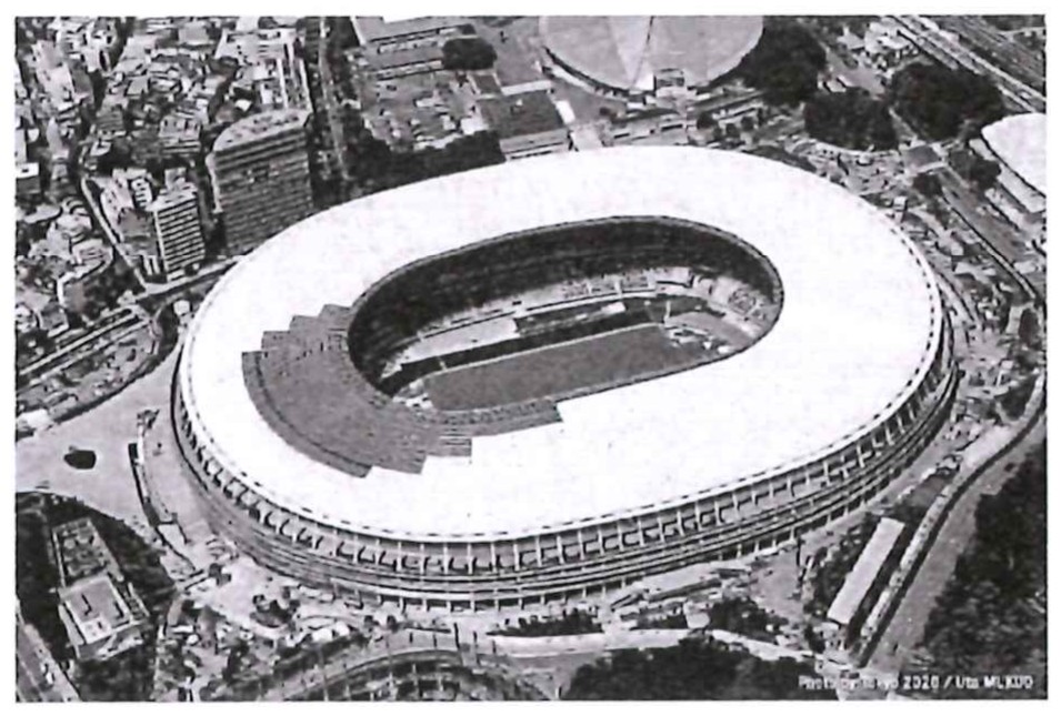 Fotografia aérea em preto e branco do Estádio Olímpico de Tóquio, de formato oval, cercado por prédios e árvores.