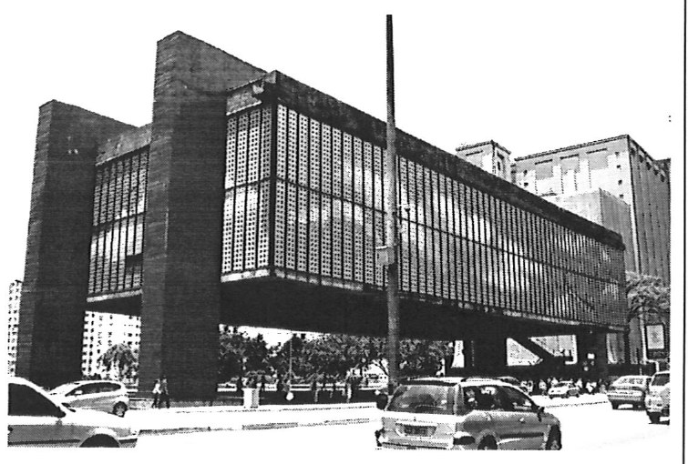 Fotografia em preto e branco de edifício com grande volume horizontal envidraçado suspenso por dois pórticos laterais de concreto, com vão livre sob o volume, em avenida com carros em primeiro plano.