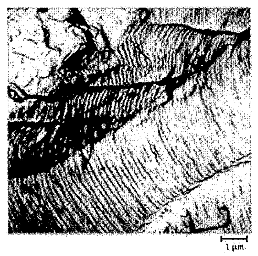 Micrografia da superfície de fratura de um alumínio com barra de escala de 1 µm, mostrando regiões claras e escuras com numerosas marcas lineares paralelas.