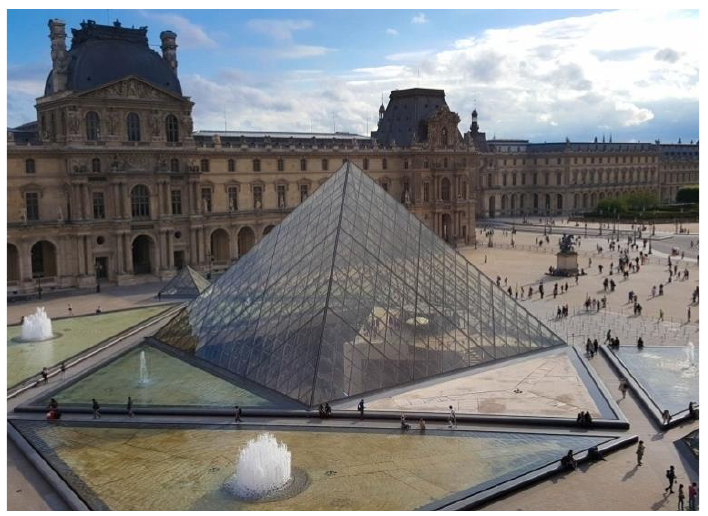 Fotografia da pirâmide de vidro do Museu do Louvre, em Paris, com o palácio ao fundo e espelhos d'água com fontes em primeiro plano.