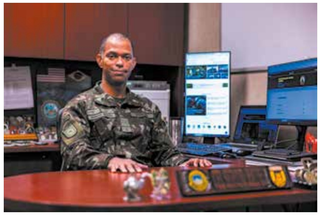 Fotografia de um militar de uniforme camuflado sentado a uma mesa de escritório, com computadores ao fundo.