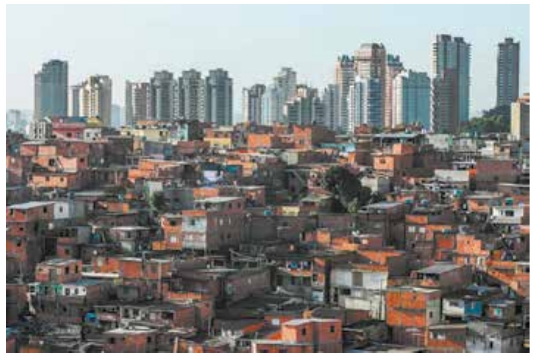 Fotografia de paisagem urbana com uma área extensa de moradias autoconstruídas de tijolos aparentes em primeiro plano e edifícios altos ao fundo.