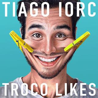 Capa do CD “Troco Likes”, de Tiago Iorc: rosto sorridente do cantor sobre fundo azul-turquesa, com dois prendedores de roupa amarelos presos às bochechas puxando o sorriso. No alto, “TIAGO IORC”; embaixo, “TROCO LIKES”.