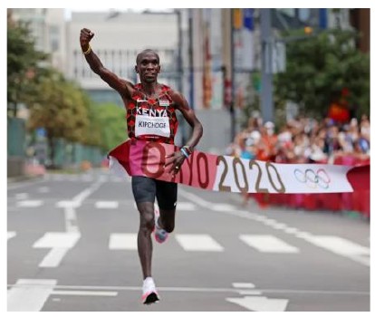 Fotografia de um maratonista queniano cruzando a faixa de chegada com o punho erguido, com a inscrição Tokyo 2020 na faixa.