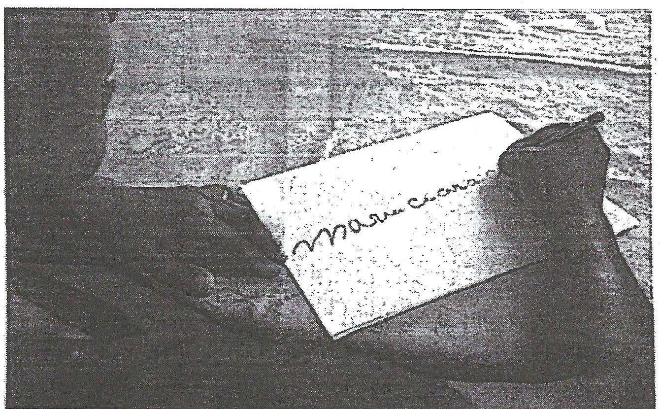 Fotografia em preto e branco de mãos de uma pessoa escrevendo à mão, com caneta, o nome “Maria Cícera” em uma folha de papel apoiada sobre uma mesa.