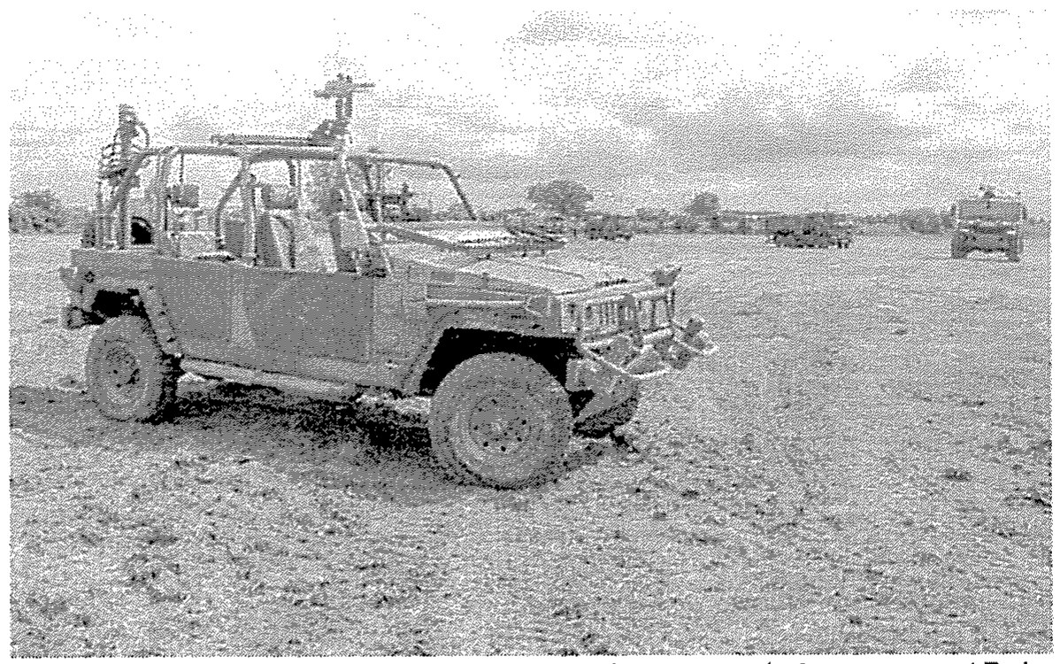 Fotografia em tons de cinza de uma viatura militar Agrale Marruá, sem capota, com arco de proteção e armamento montado, em terreno de terra; ao fundo, outras viaturas.
